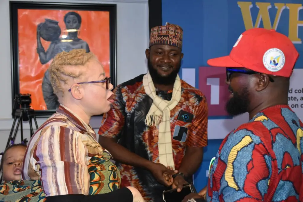 Person with albinism carrying a child engages in conversation with two men wearing traditional and casual attire at an indoor event venue