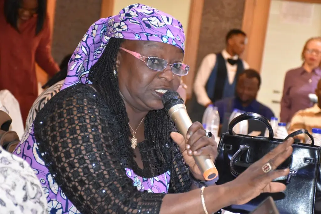Woman wearing a purple headscarf and glasses speaks into a handheld microphone while participating in a meeting, with other attendees seated in the background.