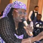 Woman wearing a purple headscarf and glasses speaks into a handheld microphone while participating in a meeting, with other attendees seated in the background.