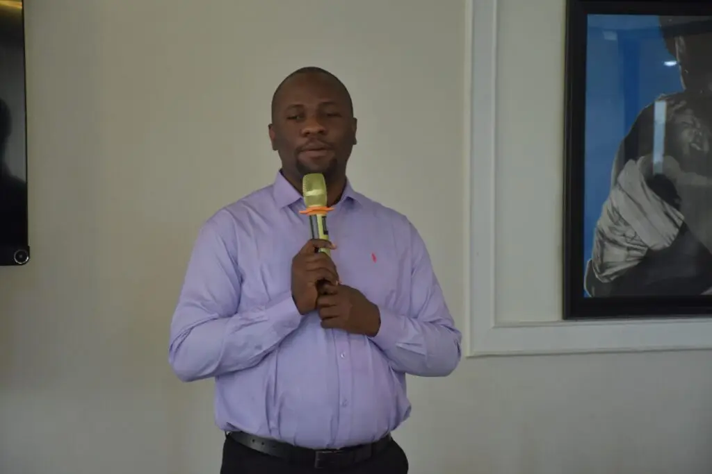 A man in a light purple shirt stands indoors holding a microphone and addressing an audience during a presentation or meeting.