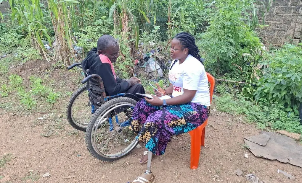 A woman seated on a plastic chair speaks with a man using a wheelchair in an outdoor rural environment. The two appear engaged in an interview or discussion, with vegetation and farmland visible in the background