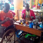 Two entrepreneurs work at a small electronics repair workstation in a busy market. One entrepreneur uses a wheelchair while handling electronic components, and another works beside a table filled with repair tools, wires, and devices. The image highlights entrepreneurship, technical skills, and economic participation among persons with disabilities.