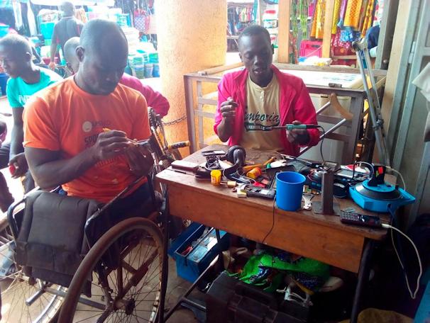 Two entrepreneurs work at a small electronics repair workstation in a busy market. One entrepreneur uses a wheelchair while handling electronic components, and another works beside a table filled with repair tools, wires, and devices. The image highlights entrepreneurship, technical skills, and economic participation among persons with disabilities.